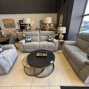 A furniture showroom displays the Barrington Electric Recliner with supportive cushioning and floral pillows, a round black coffee table, and table lamps. Large windows provide natural light, with framed artwork on the back wall.
