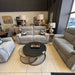 A furniture showroom displays the Barrington Electric Recliner with supportive cushioning and floral pillows, a round black coffee table, and table lamps. Large windows provide natural light, with framed artwork on the back wall.