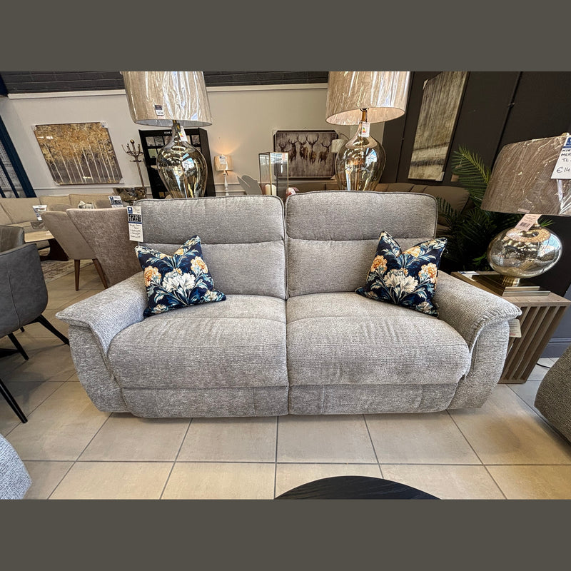 The Barrington Electric Recliner, a modern light gray fabric seat with durable upholstery and two floral-patterned throw pillows, is displayed in a showroom among side tables, lamps, and other furniture pieces.