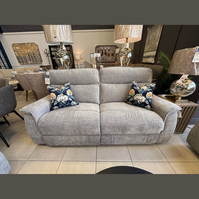 The Barrington Electric Recliner, a modern light gray fabric seat with durable upholstery and two floral-patterned throw pillows, is displayed in a showroom among side tables, lamps, and other furniture pieces.