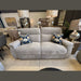 The Barrington Electric Recliner, a modern light gray fabric seat with durable upholstery and two floral-patterned throw pillows, is displayed in a showroom among side tables, lamps, and other furniture pieces.