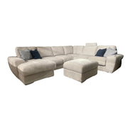 The Grand Corner Group 4L is a large beige modular sofa with a left chaise, matching ottoman, and assorted dark and light throw pillows, set against a white background. It features micro pocket springs for enhanced comfort.