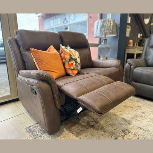 The Logan Recliner Sofa, a brown leather loveseat with three colorful throw pillows—orange and floral—sits in a showroom near a window, paired with a lamp on a side table.