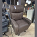 The Hugo Lift & Rise, a large brown reclining armchair with twin motors and leg support, is displayed in a furniture store on a tiled floor beside a dark gray chair, with a price tag attached.