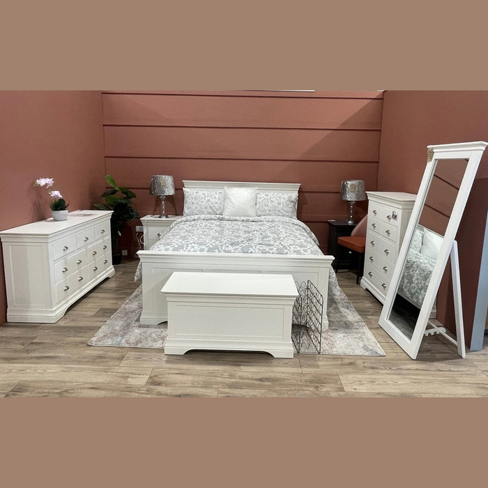 A bedroom featuring the Bella Bedframe in white, paired with matching nightstands, dressers with chrome cup handles, and a bench. The gray patterned comforter, large mirror, plants, lamps, and soft rug complete the space.