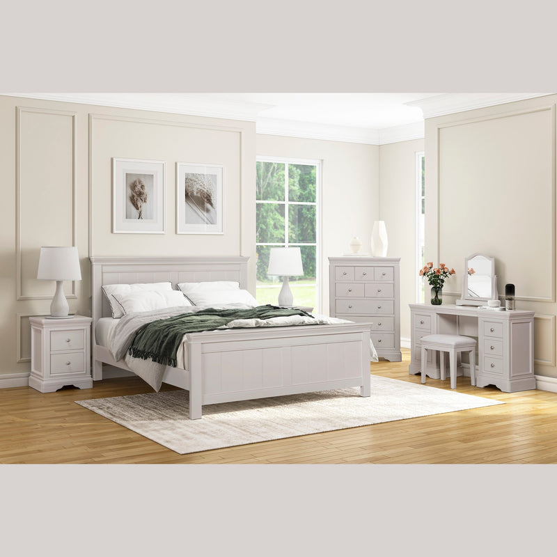 A bright, airy bedroom highlights the Lima Tall Chest from the Lima Taupe Bedroom Range, paired with a taupe bed frame, white nightstands, dresser, and vanity. Light bedding, a green throw, lamps, and artwork enhance the space as sunlight pours in.