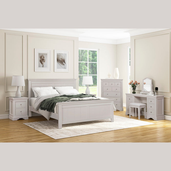A bright, modern bedroom with white furniture, including a bed, bedside tables, the Lima Wide Chest, vanity, and stool. Large windows brighten the space; framed art adorns walls and a rug highlights the wooden floor.