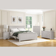 A bright bedroom features the Lima Tall Chest along with white and taupe furniture, a bed, and nightstands. Large windows fill the room with light; lamps, photos, and fresh flowers add decorative touches.
