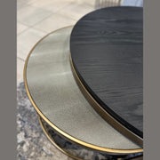 Close-up of the Noir Coffee Table Nest: one table has a textured light metallic top, the other a smooth dark wood surface; both feature gold-colored rims and rest on a gray carpeted floor.
