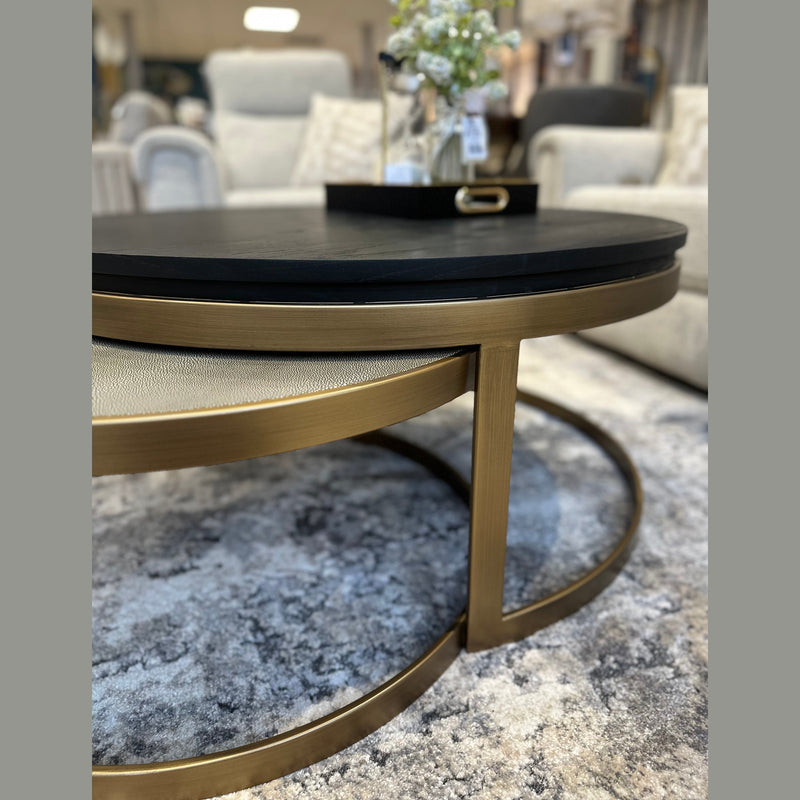 A close-up of the Noir Coffee Table Nest featuring a sleek black top and gold metal frame on a patterned rug, with a tray of flowers atop it and neutral sofas in the background.