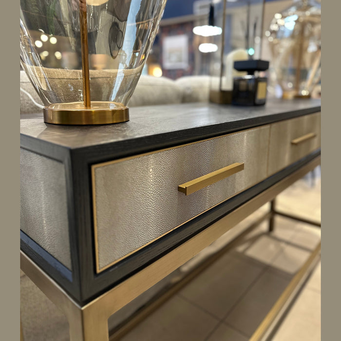 A close-up of the Noir Console, a luxury table with a textured metallic drawer front and gold handle, topped with a glass lamp with a gold base, showcased in a stylish interior setting.