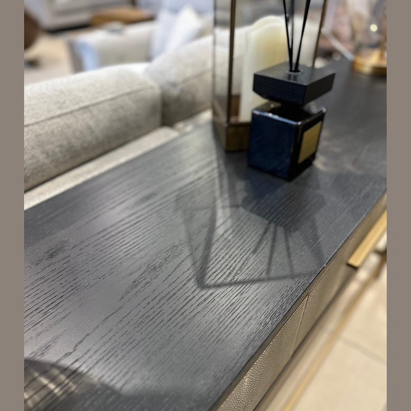 A close-up captures the Noir Console with gold handles, featuring modern decor—a candle and diffuser—on its dark wood surface, while a blurred sofa and cushions appear in the background.