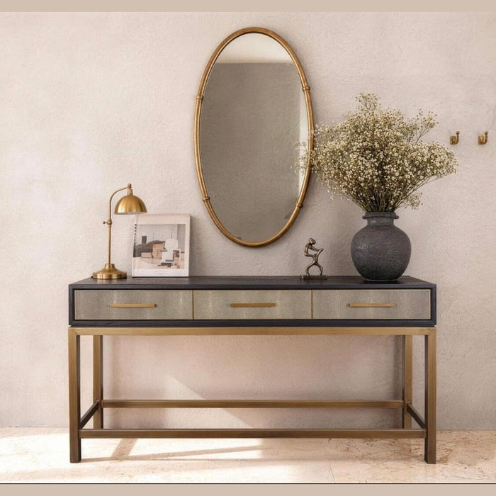 The Noir Console, a modern dark wood piece with gold accents and three drawers, is styled with a gold lamp, framed art, small figurine, large vase of white flowers. Above it hang an oval mirror and wall hooks on a light-toned wall.