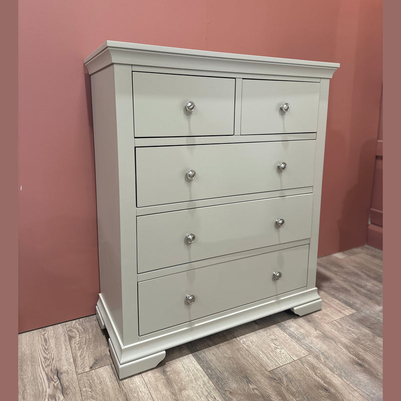 The Malmo Tall Chest Of Drawers in light gray has 5 drawers, round metal knobs, and a slightly curved base. Its modern, simple design enhances bedroom storage and looks stylish on a wood floor against a pink wall.