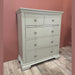 The Malmo Tall Chest Of Drawers in light gray has 5 drawers, round metal knobs, and a slightly curved base. Its modern, simple design enhances bedroom storage and looks stylish on a wood floor against a pink wall.