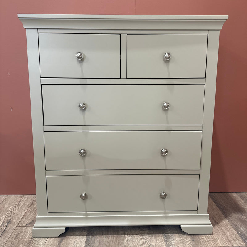 The Malmo Tall Chest Of Drawers in cream features two small top drawers and three larger ones with metal knobs, offering ample bedroom storage. It stands on a wooden floor against a reddish-brown wall.