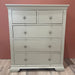 The Malmo Tall Chest Of Drawers in cream features two small top drawers and three larger ones with metal knobs, offering ample bedroom storage. It stands on a wooden floor against a reddish-brown wall.