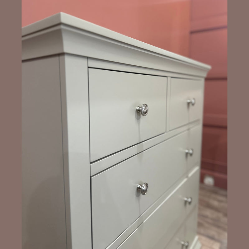 A close-up of the Malmo Tall Chest Of Drawers, a white wooden unit with four drawers and metal knobs, set against a pink wall and hardwood floor—ideal for adding stylish storage to any bedroom.