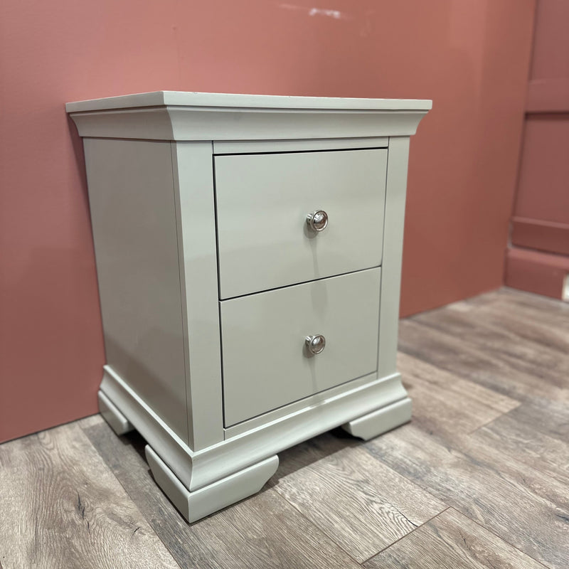 The Malmo Locker, a light gray bedside table with two drawers and silver knobs, provides durable storage on wood flooring against a muted pink wall.
