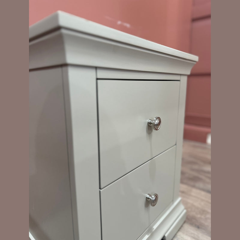 A close-up of the Malmo Locker, a white two-drawer nightstand with silver knobs, offers durable storage. It stands against a pink wall and rests on a wooden floor.