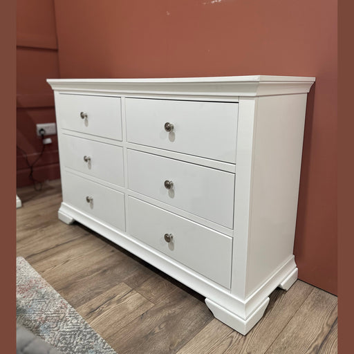 Part of the Malmo Bedroom Collection, the Malmo Wide Chest of Drawers, finished in white with silver knobs, stands on a wood floor against a reddish-brown wall; a mattress and an electrical outlet appear nearby.