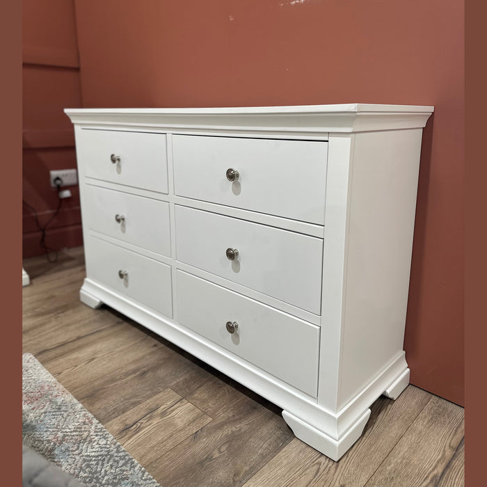 Part of the Malmo Bedroom Collection, the Malmo Wide Chest of Drawers, finished in white with silver knobs, stands on a wood floor against a reddish-brown wall; a mattress and an electrical outlet appear nearby.