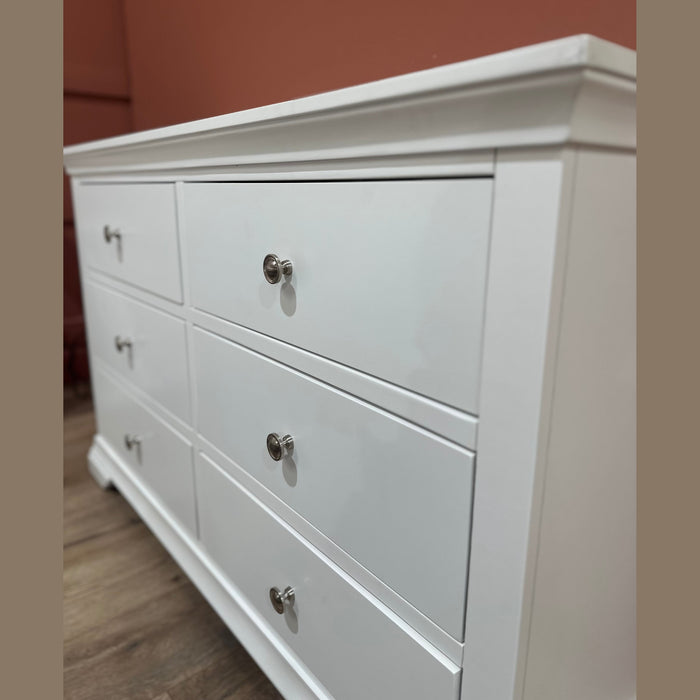 A white Malmo Wide Chest of Drawers with six drawers and silver knobs stands on a wooden floor against a reddish-brown wall. The angled photo showcases its elegant design from both the side and front.