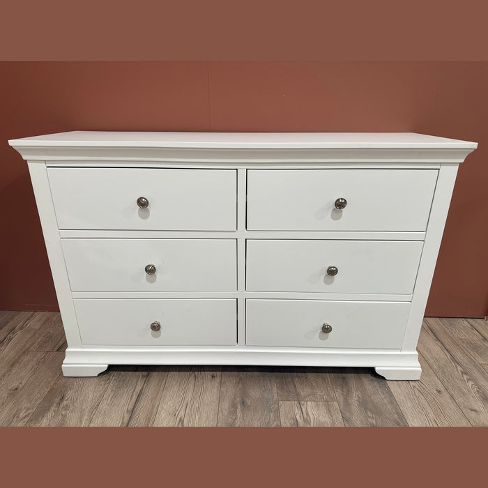 The Malmo Wide Chest of Drawers, with six white drawers, silver knobs, and a raised base, stands on a wooden floor against a reddish-brown wall—an ideal choice for modern white bedroom decor.