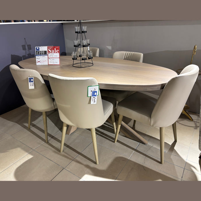 A light wood oval dining table is surrounded by six Patrice Dining Chairs with taupe leather cushions and sale tags, displayed in a tiled showroom. A tiered candle holder sits at the center of the table.