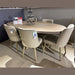 A light wood oval dining table is surrounded by six Patrice Dining Chairs with taupe leather cushions and sale tags, displayed in a tiled showroom. A tiered candle holder sits at the center of the table.