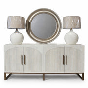 The Marvel Sideboard features modern fluted doors with vertical grooves and gold handles, topped with two plastic-wrapped white lamps and a large round silver-framed mirror centered above.