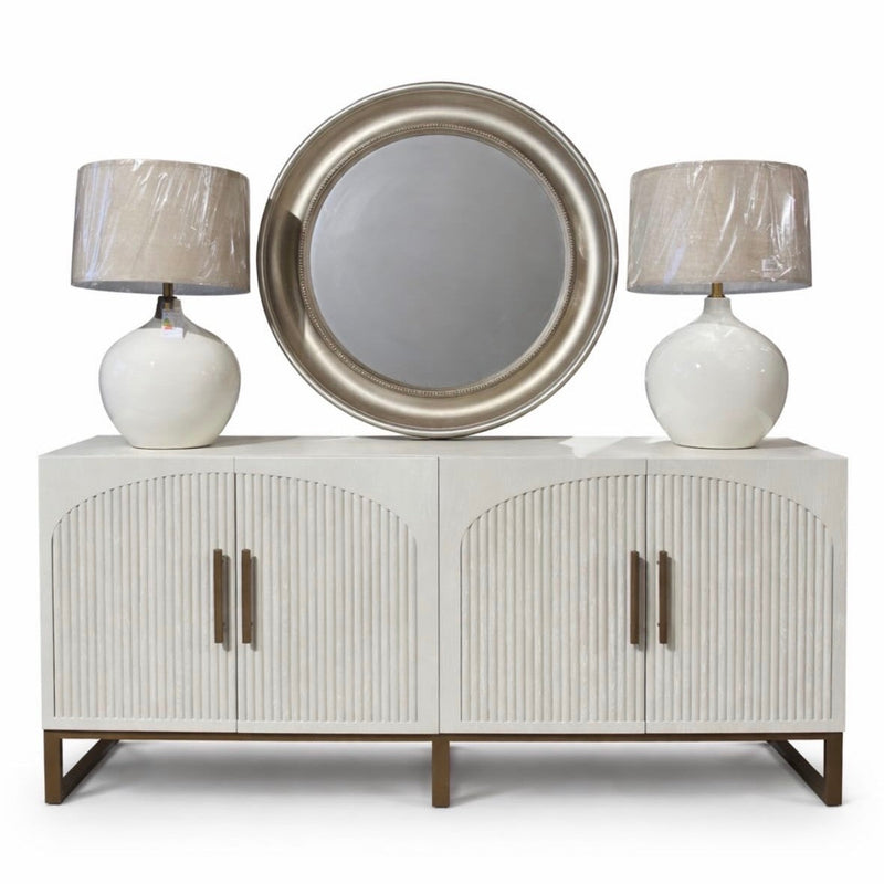 The Marvel Sideboard features modern fluted doors with vertical grooves and gold handles, topped with two plastic-wrapped white lamps and a large round silver-framed mirror centered above.