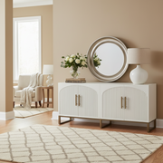 A bright, modern living room showcases the Marvel Sideboard with gold handles and fluted doors, topped with a round mirror, lamp, and flowers. A cream armchair, soft throw, and geometric rug complete the stylish neutral décor.