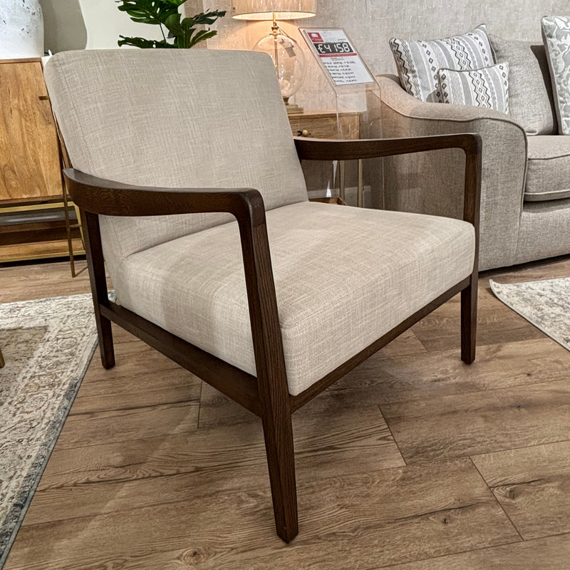 The Monza Armchair, featuring a walnut wood frame and light beige upholstered seat and backrest, is showcased on a wooden floor in a stylish living room display.