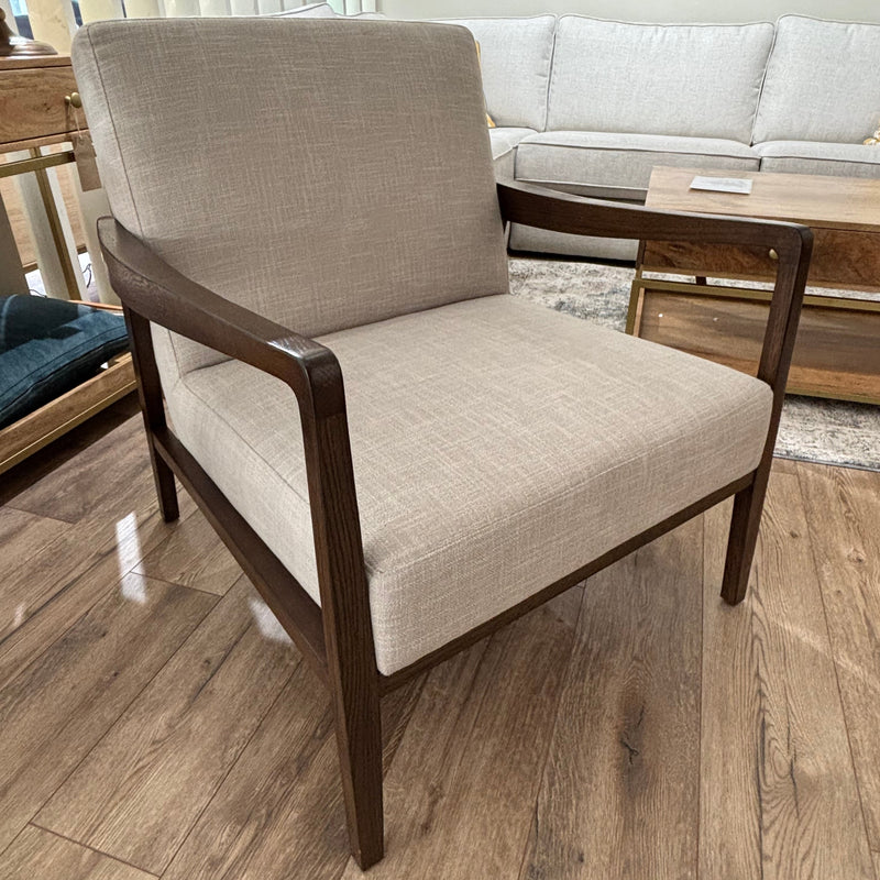 The Monza Armchair, featuring beige linen and a walnut wood frame, sits on a wooden floor in the living room near a light sofa, coffee table, and window with vertical blinds.