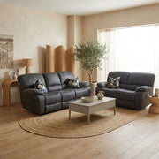 A cozy living room featuring the Sapphire Reclining Sofa in dark gray with floral pillows, a light coffee table, a round woven rug, wood decor accents, a potted olive tree, and large windows welcoming natural light.