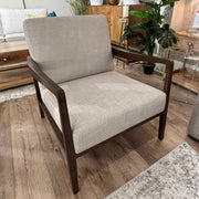 The Monza Armchair, featuring beige linen fabric and a dark walnut wood frame, sits on a wooden floor beside indoor plants, cabinets, and decor accessories in a modern living room setting.