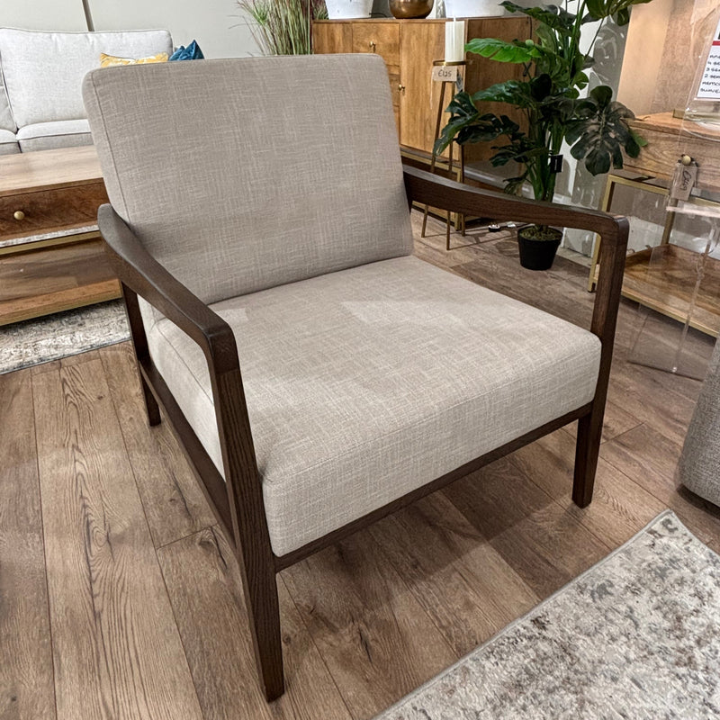 The Monza Armchair, featuring beige linen fabric and a dark walnut wood frame, sits on a wooden floor beside indoor plants, cabinets, and decor accessories in a modern living room setting.