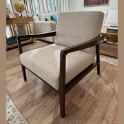 The Monza Armchair, upholstered in beige linen with a dark walnut wood frame, sits on a wooden floor in a modern living room featuring a sofa, coffee table, vertical blinds, and a horse painting on the wall.