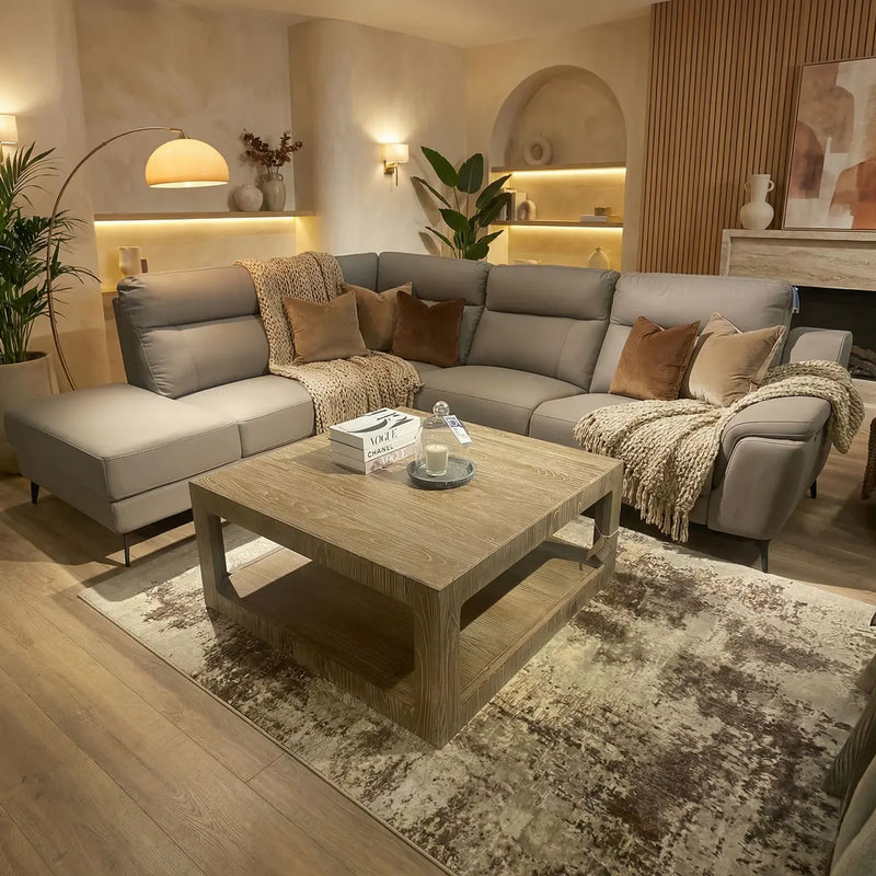 A cozy living room with the modern Chloe Corner Group electric reclining sofa in taupe, brown and beige cushions, a wood coffee table, decor accents, a patterned rug, plants, soft lighting, neutral walls, and built-in shelves.