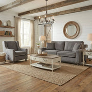 A cozy living room featuring the Emperor Range gray Irish-made sofa and armchair in Aqua Clean fabrics, with white coffee and side tables, round mirror, rustic beams, jute rug, neutral decor, and sunlight streaming through beige curtains.