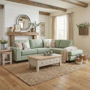 A cozy living room with the Harold Chaise Corner sofa in light green, pastel floral pillows, a wooden coffee table, woven rug, rustic decor like a round mirror, framed art, and baskets on cream walls and wood floors.