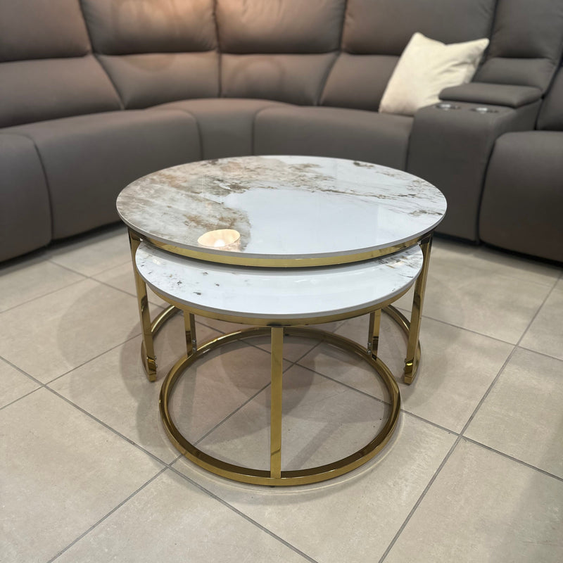 The Kas Nest - Gold Base, a set of two round nesting coffee tables with white marble tops and gold frames, stands on a tiled floor before a gray sectional sofa with a cream pillow, complemented by a lamp and wall art.