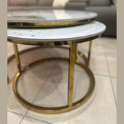 Two Kas Nest - Gold Base round modern nesting coffee tables with white marble tops and gold frames sit on a tiled floor in front of a dark gray sofa with light-colored cushions.
