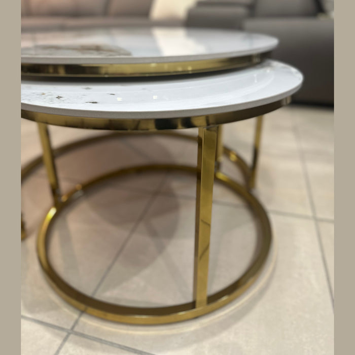 Two Kas Nest - Gold Base round modern nesting coffee tables with white marble tops and gold frames sit on a tiled floor in front of a dark gray sofa with light-colored cushions.