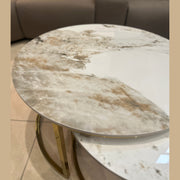 The Kas Nest - Gold Base, a modern round coffee table with a white and light brown marbled surface and gold legs, sits on a tiled floor in front of a brown sofa adorned with beige and white cushions, adding elegance to the room.
