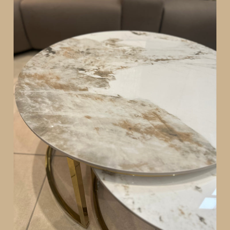 The Kas Nest - Gold Base, a modern round coffee table with a white and light brown marbled surface and gold legs, sits on a tiled floor in front of a brown sofa adorned with beige and white cushions, adding elegance to the room.