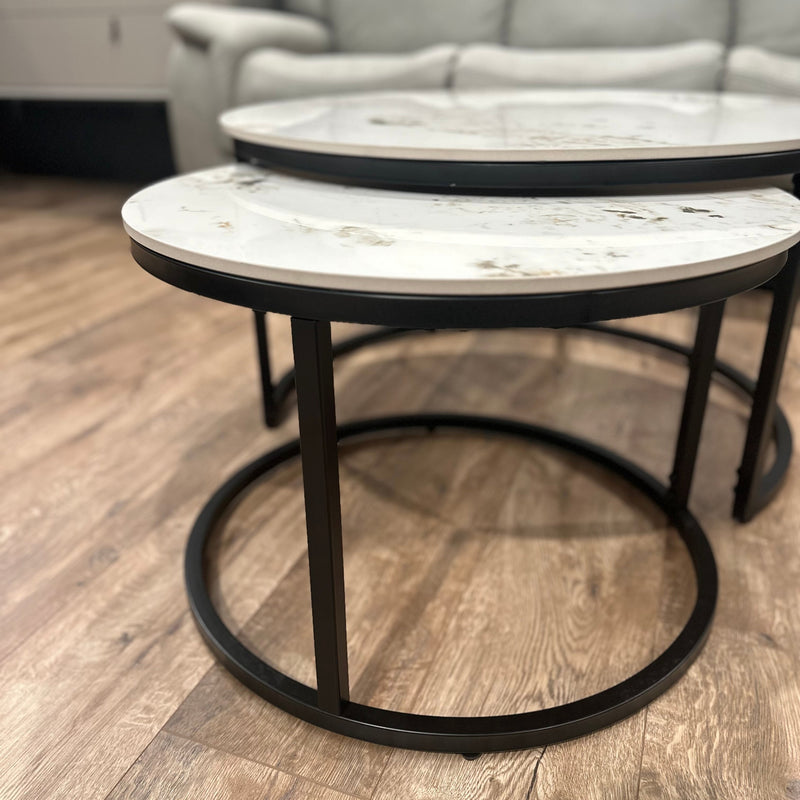 A close-up of Kas Nest, two round nesting coffee tables featuring white marble tops and black metal frames on a wooden floor. A light-colored sofa and a cabinet complete the modern background.