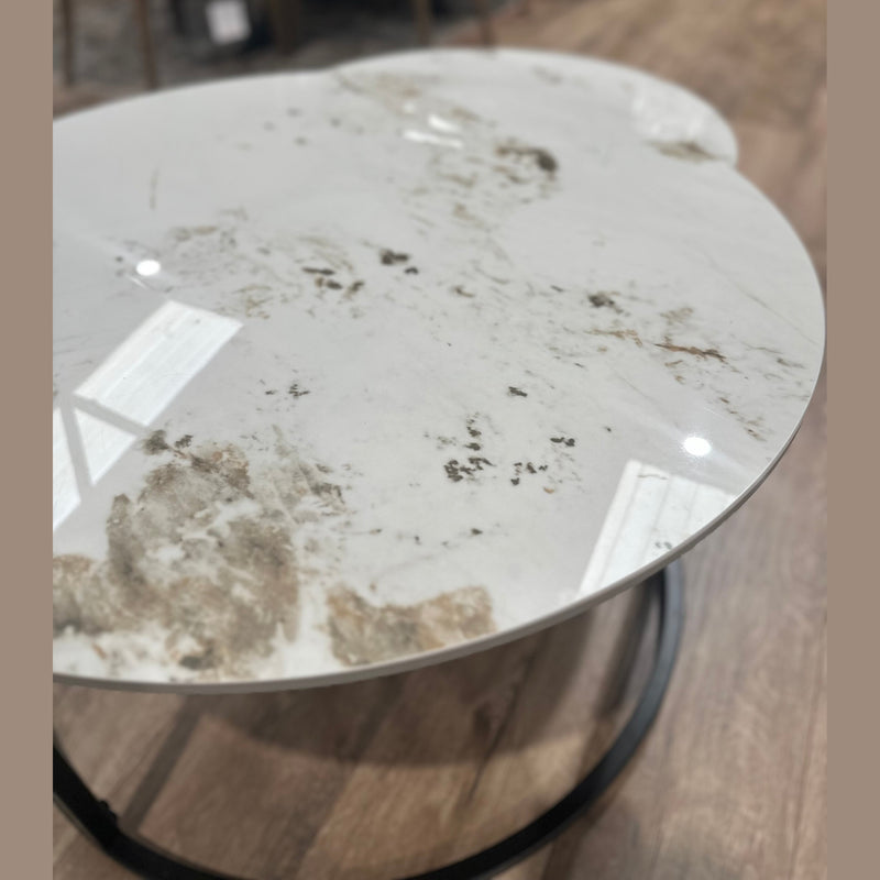 The Kas Nest coffee table features a round white marble top with brown and gray veining, reflecting ceiling lights, on a dark metal base set atop a wooden floor. A blurred modern dining area is visible in the background.