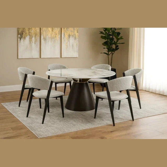 A round dining table with a dark conical base is paired with six Titan Dining Chairs in light gray stone fabric on a textured rug. Abstract artwork and a tall potted plant accent the neutral-toned room.